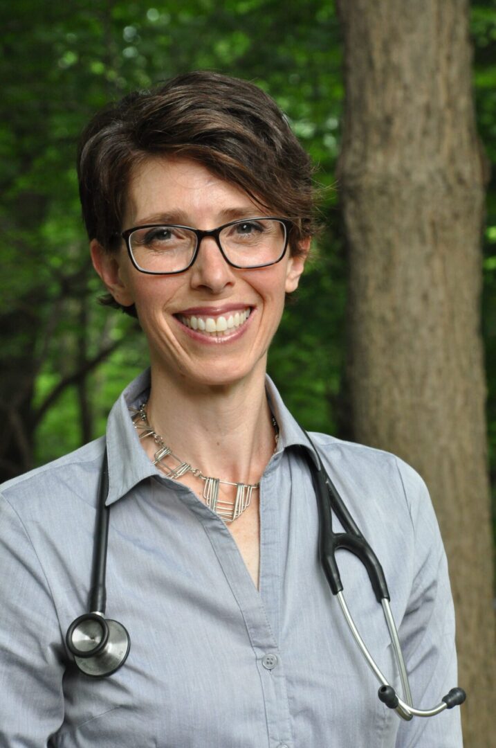 A woman with glasses and stethoscope standing in front of trees.
